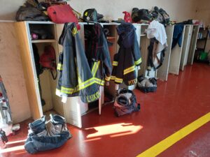 Fire department locker room. Firefighters' apparel is strewn about, jackets draped over open locker doors.