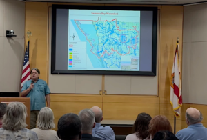 Steve Suau stands with a microphone in front of an audience. Behind him is a projected map titled Sarasota Bay Watershed.