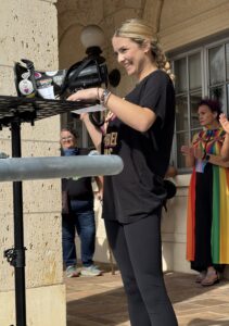 Ava Donoghue speaks into a megaphone adorned with LGBTQIA+ stickers and one that reads "Rise Up."