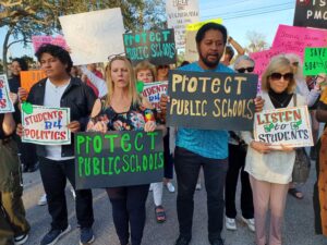 Protesters hold signs supporting education with phrases like "Protect public schools," "listen to students," "students B4 politics," and "fund DOE and fire DOGE."