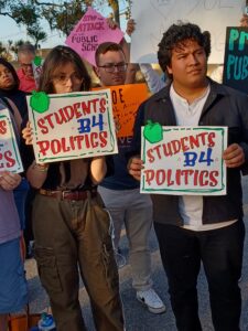 Two protestors hold up identical signs that say "Students B4 Politics".