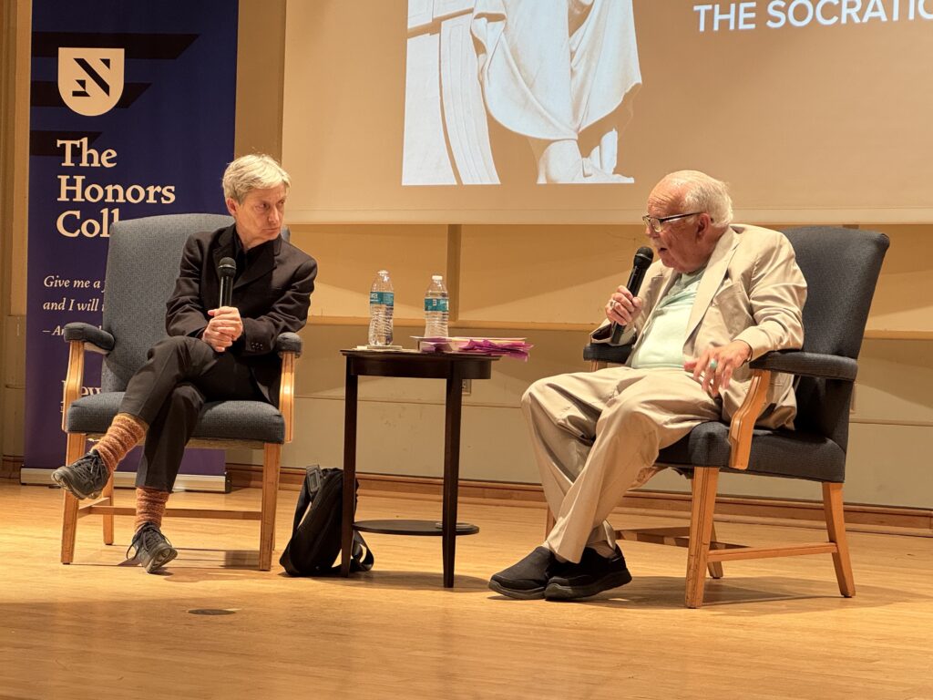 Judith Butler and Stanley Fish sitting and talking on stage holding microphones.