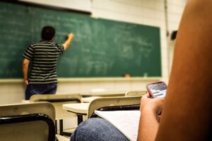 Over-the-shoulder photo in a classroom of a student on their cellphone while a teacher with their back to the student writes on a chalkboard.