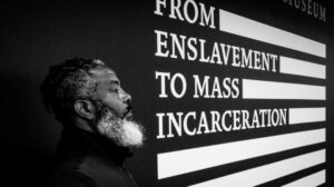 Black and white photo of a bearded Black person facing to the right toward all-caps text on the wall beside them which reads "From enslavement to mass incarceration."