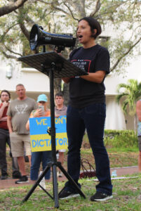 A speaker with a megaphone addresses a crowd. Behind them, a protester holds a sign with the Ukrainian flag colors which reads "We don't do dictators."