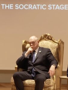 Tom Homan sitting in an ornate golden chair on stage with the text "The Socratic Stage" visible above him.