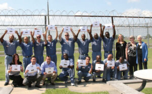 Individuals smile and hold up certificates in front of a fence topped with barbed wire.