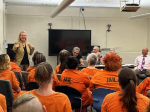 Theresa Cusimano addresses a small crowd of people wearing orange jumpsuits that say "jail" on the back.