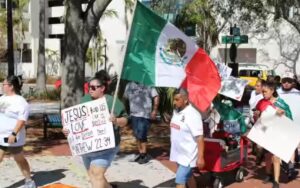 Pro-immigrant protestors walk by, one of them holding a Mexican flag and a sign that reads "Jesus told us to love our neighbors, not to deport them! Matthew 22:34".