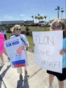 Two protesters holding signs that say "Elon, DOGE yourself!" and "No one voted for Elon Musk".