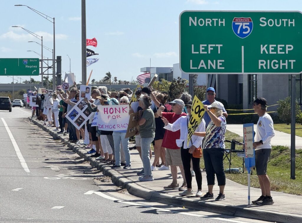 A long line of anti-Musk protesters on the sidewalk along University Parkway. One of the protesters' signs reads "Honk for democracy".