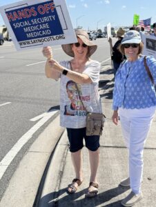 Photo of two protesters. One cheerfully holds up a sign that reads "Hands off Social Security, Medicare, Medicaid" and "www.saynocuts.org".