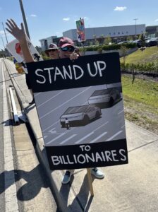 A protester holds up a sign that reads "Stand up to billionaires" with an illustration of a person standing in the road blocking three approaching Tesla Cybertrucks.