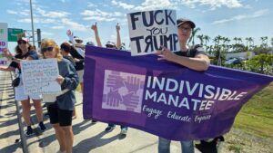 Protesters wave signs including one that reads "Fuck you Trump" (with a backwards "R") and hold a banner with the logo and name of the organization Indivisable Manatee.