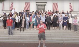 Dozens of people assemble on the steps outside a building while a speaker front and center with a microphone addresses the viewer.