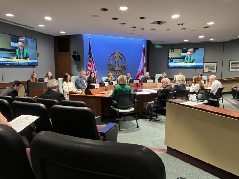 A 13-member committee sits around a conference table. the City of Sarasota logo is on the wall behind them.