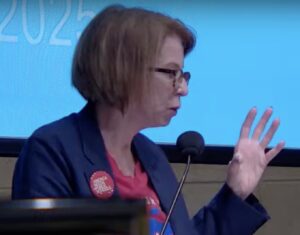 Lisa Schurr, wearing an "America Needs Public Schools" button, speaks into a microphone.