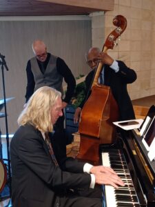 A pianist plays in the foreground. A standup bassist plays behind.