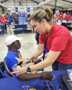 In a large, open building with a crowd in the background, a person with a stethoscope checks a patient's blood pressure.