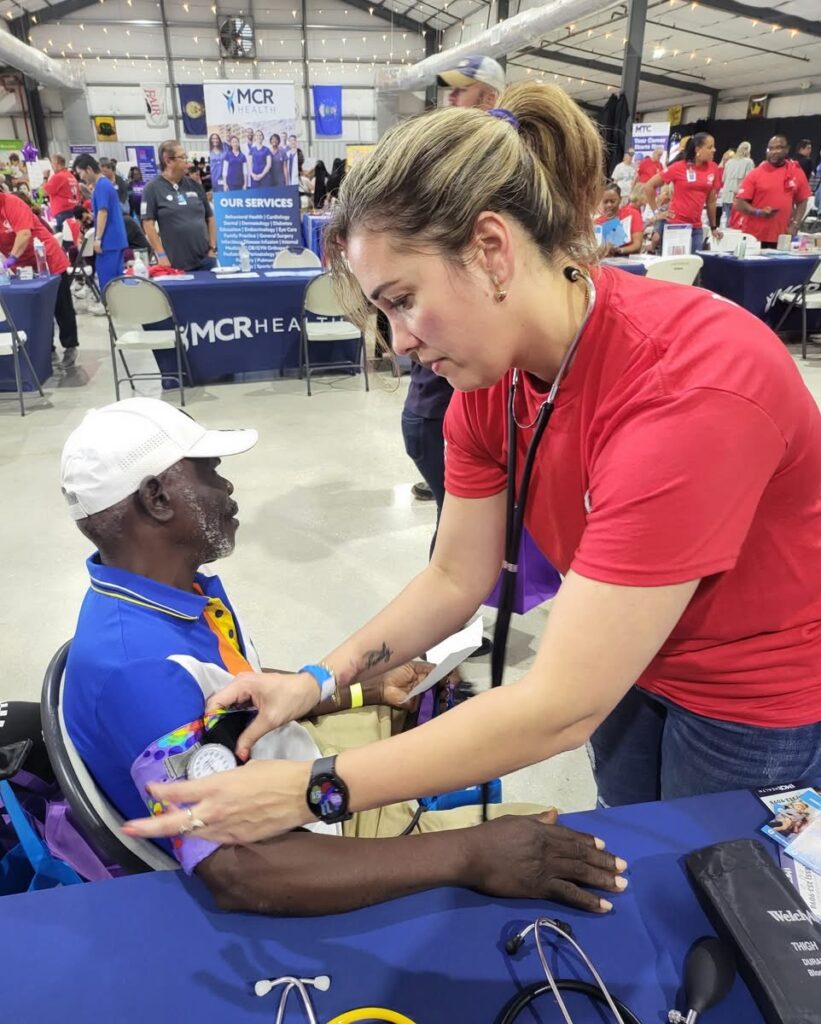 In a large, open building with a crowd in the background, a person with a stethoscope checks a patient's blood pressure.