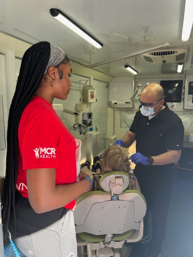 A dentist examines a patient's teeth. An employee in the foreground wears a shirt that says "MCR Health" on the sleeve.
