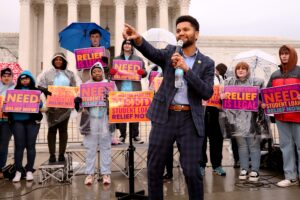 Max Frost speaking into a microphone and pointing toward the crowd. Demonstrators behind Frost hold signs in support of student loan relief.