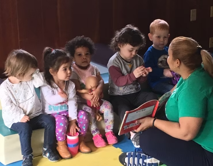 Five children sit attentively—though one looks at the camera—as an adult reads them a picture book.