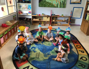 Children wearing paper animal and character hats sit on the floor of a classroom on a world map rug, looking at the camera.