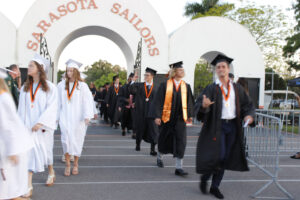 A procession of high school graduates passes under an archway with lettering that reads "Sarasota Sailors."