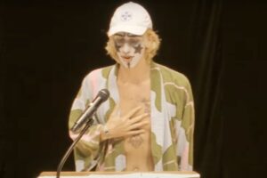Safari Svensson wearing black and white face makeup and an open front top behind a podium speaking.