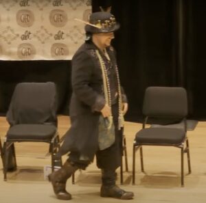 A student on stage wearing an elaborate costume including a long overcoat, leather boots and a pork pie hat.