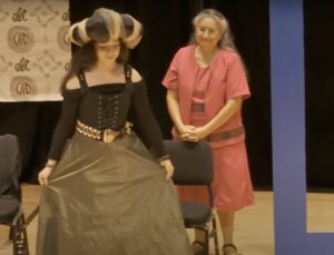 A student wearing a long skirt, a corset, and an elaborate hat curtsies on stage.