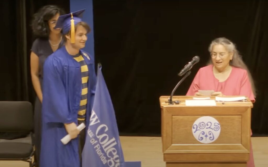 A student wearing a blue cap and gown and carrying a New College flag approaches a speaker behind a podium.