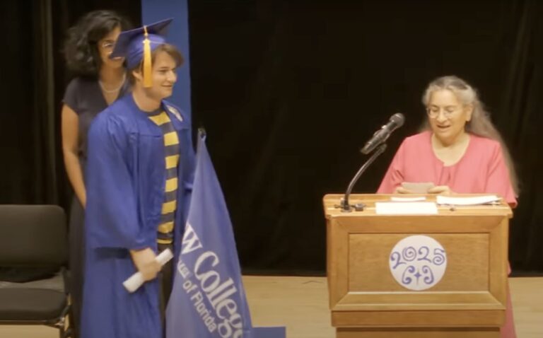 A student wearing a blue cap and gown and carrying a New College flag approaches a speaker behind a podium.