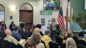 A speaker addresses a crowd at an official venue. Signage indicates this is photo is of a sustainable communities workshop on November 7, 2024.
