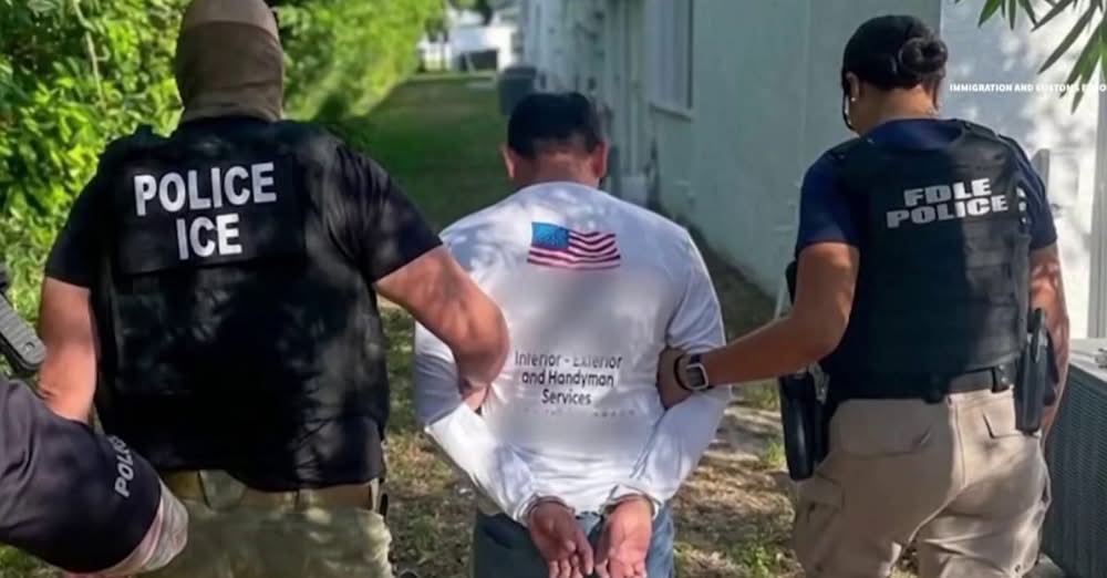 Two officers, one of ICE and one of the FDLE police, escort a cuffed detainee. The back of the detainee's shirt is mostly blurred, but the text "Interior - Exterior and Handyman Services" is legible.