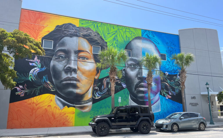 A huge mural depicting Irene and Rev. Lewis Colson on the wall of a building. Abstract floral patterns fill the colorful background. Half of each subject's face is in greyscale.