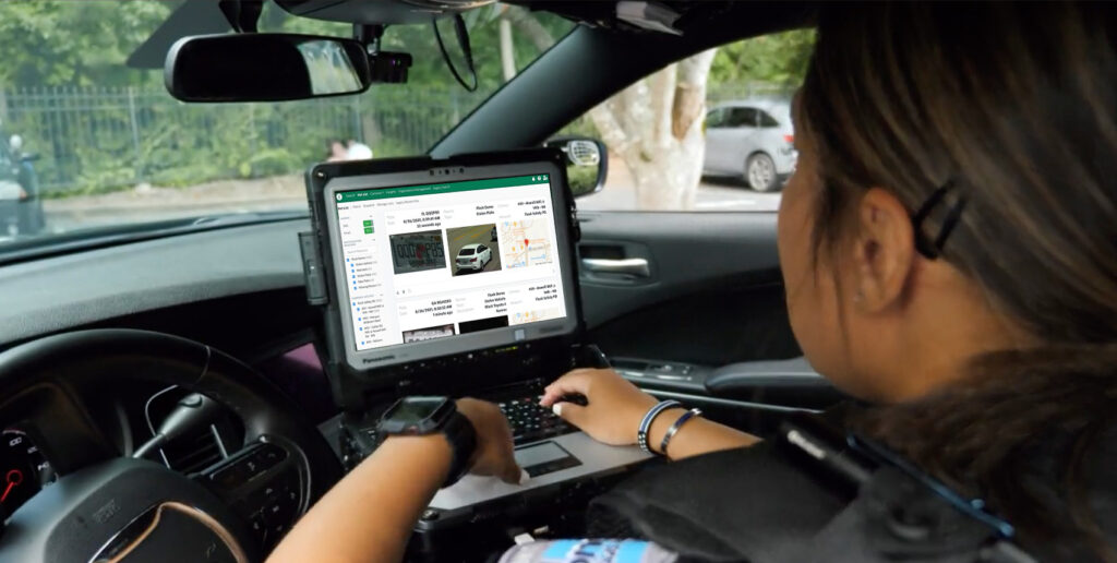 Over-the-shoulder photo of a police officer in a patrol car viewing license plate data on a laptop.