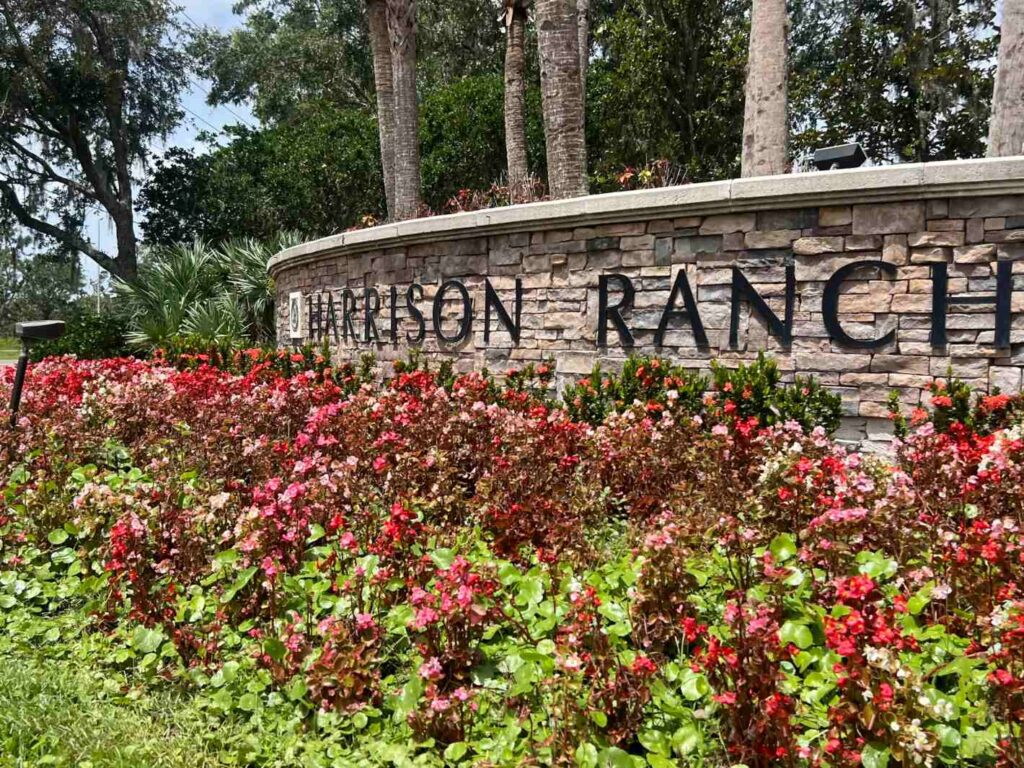 Mixed stone sign that reads "Harrison Ranch" in front of palm trees and behind landscaped foliage.