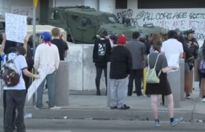 Shot of a group of protesters. One holds a sign that reads "soy la voz de mis padres." Graffiti spray painted on a wall appears to read "All cops are bitches."