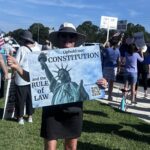 A demonstrator at a protest holds a sign that says "Uphold our constitution and the rule of law" with a photo of the Statue of Liberty." In the background, another demonstrator's handwritten sign reads "The Turd Reich."