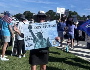 A demonstrator at a protest holds a sign that says "Uphold our constitution and the rule of law" with a photo of the Statue of Liberty." In the background, another demonstrator's handwritten sign reads "The Turd Reich."