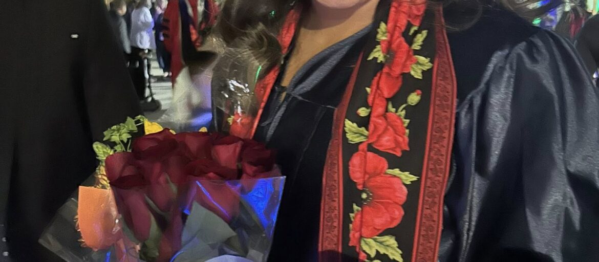 Maria Martinez in her cap and gown holding a rose bouquet while smiling at her phone.
