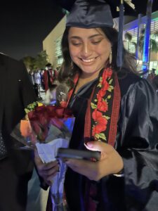 Maria Martinez in her cap and gown holding a rose bouquet while smiling at her phone.