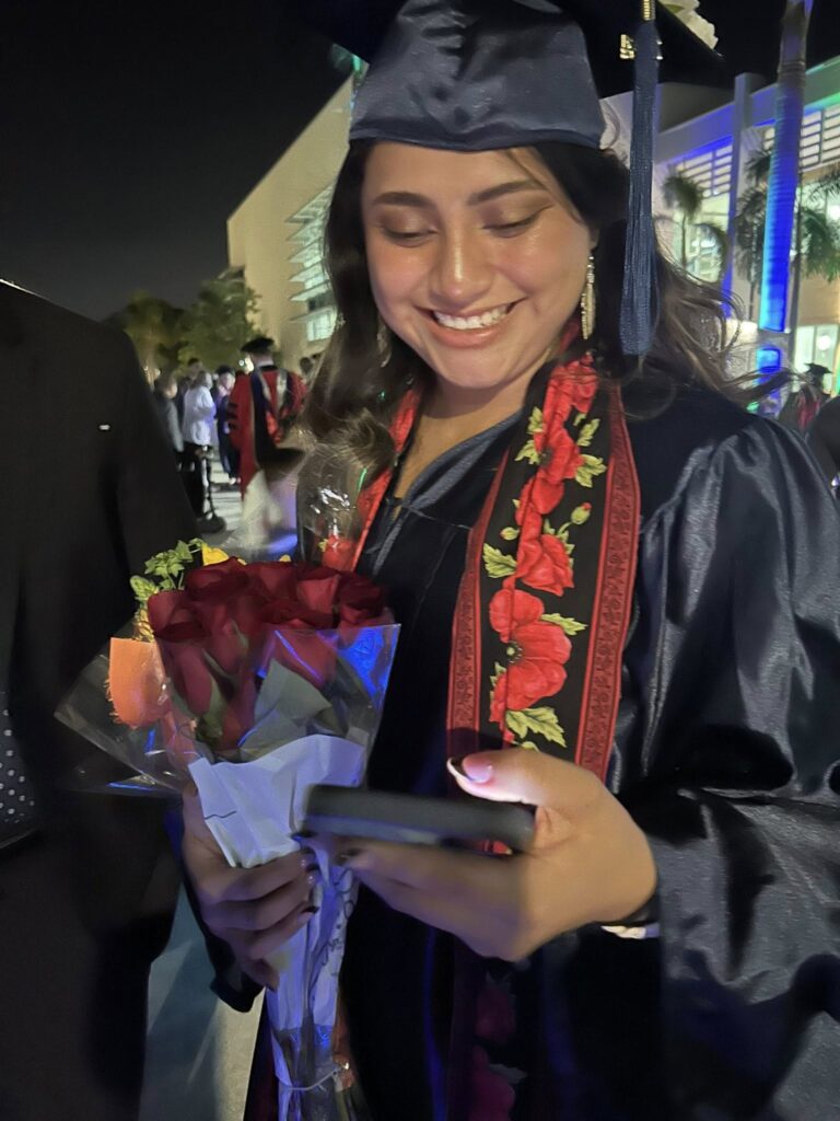 Maria Martinez in her cap and gown holding a rose bouquet while smiling at her phone.
