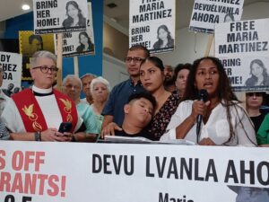 A crowd stands behind a speaker, many holding signs that read "Free Maria Martinez / Bring her home!" and "Devuélvanla ahora".