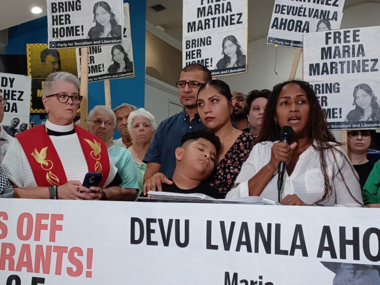 A crowd stands behind a speaker, many holding signs that read "Free Maria Martinez / Bring her home!" and "Devuélvanla ahora".