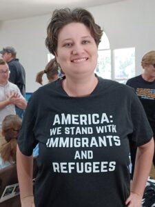 A smiling person wearing a t-shirt that reads, "America: We stand with immigrants and refugees."
