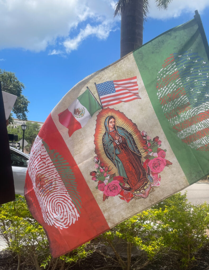 Photo of a Mexican tricolor flag with the Lady of Guadalupe with a U.S. and Mexican flags waving behind her taking the place of the national coat of arms on the central white stripe. On the green stripe is a fingerprint with the U.S. flag visible in the ink; on the red stripe is a fingerprint with the Mexican flag visible in the ink.