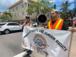 Two demonstrators, one of whom holds a megaphone, carry a banner that reads "Immigrant rights are human rights" with the logo of El Pueblo Unido Tampa Bay.